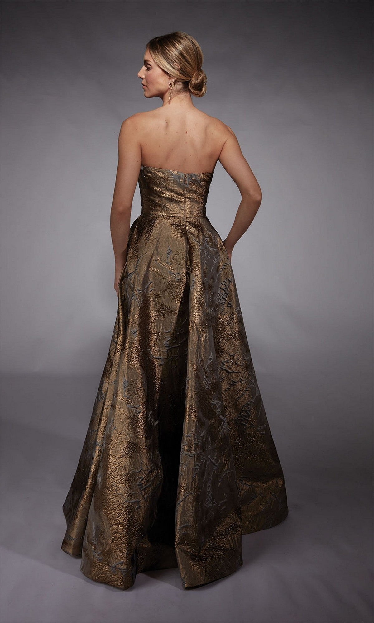 A woman stands with her back to the camera against a gray background, wearing the Alyce 27760 long formal dress—a strapless jacquard gown with a metallic gold finish.